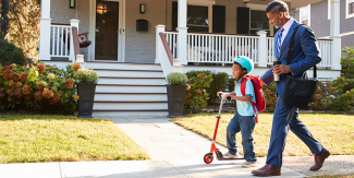 Child riding a scooter in front of a house with their parent walking closely behind
