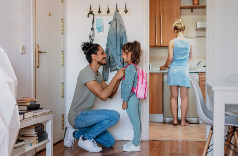 A father helps his daughter with her backpack in a cozy home entrance, while a woman prepares a meal in the background.