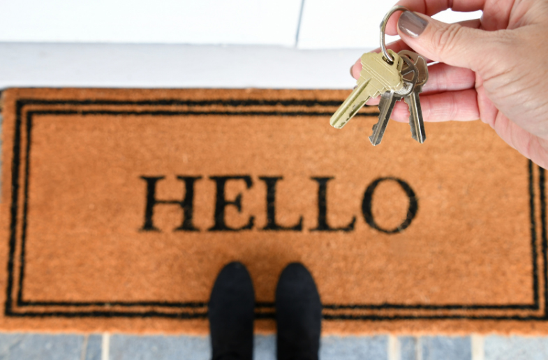 Close-up of a person's hand holding keys above a doormat that says Hello, with the person's feet visible in black shoes.