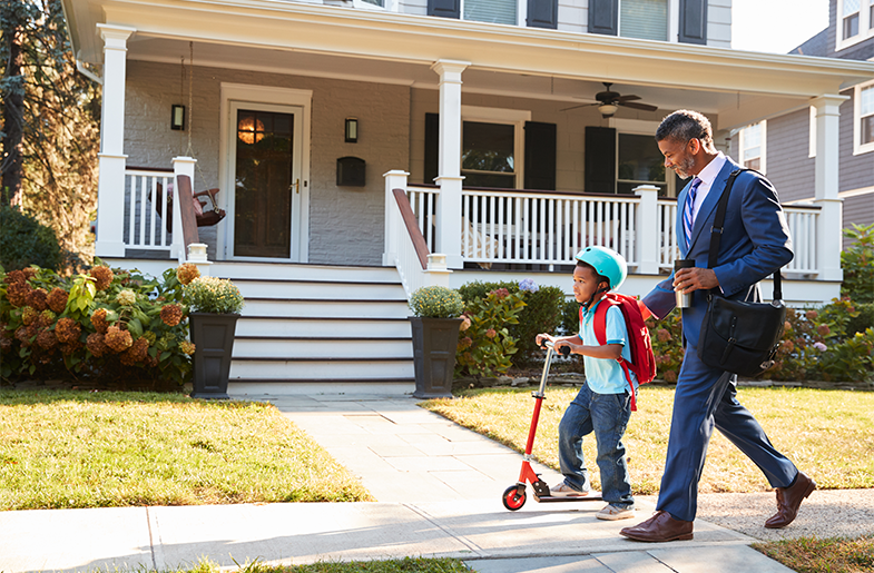 Child riding a scooter in front of a house with their parent walking closely behind