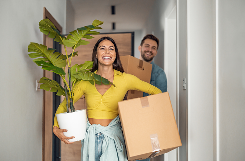Woman carries plant and box into new housing space accompanied by a friend.