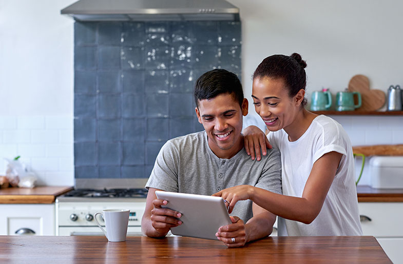 A couple is sitting closely at a kitchen counter, smiling and looking at an iPad screen