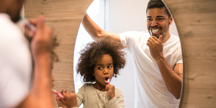 Father and daughter brush teeth together in a bathroom mirror