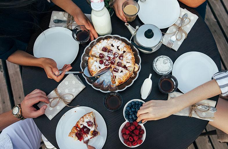 A dining table laid out with place settings, pie, berries, and coffee. Hands reaching in to enjoy.