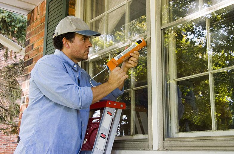 Man on a ladder using a caulk gun to caulk window trim.
